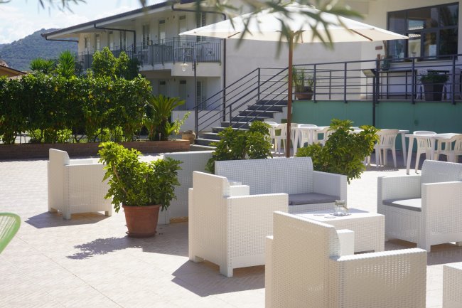 Terrazza con sedie bianche, tavoli e vegetazione, palazzo bianco sullo sfondo.
