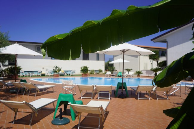 Piscina con lettini e ombrelloni, visibili foglie di banano.