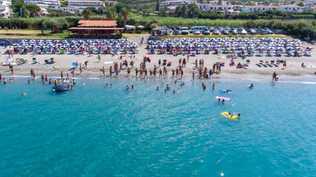 Spiaggia affollata con persone in acqua, ombrelloni in fila e chiosco.
