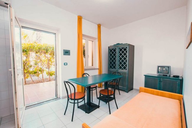 Area soggiorno con tavolo blu, sedie nere, armadio, letto singolo e porte aperte su giardino.