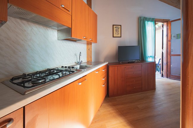 Cucina moderna con armadi in legno, piano cottura e lavandino. TV su mobile e porta-finestra aperta.