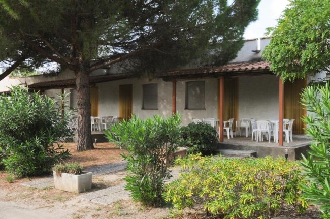 Una fila di bungalow con verande, tavoli bianchi e vegetazione circostante.