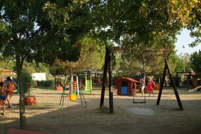 Un parco giochi all'aperto con altalene, casetta rossa e verde e scivolo, circondato da alberi.