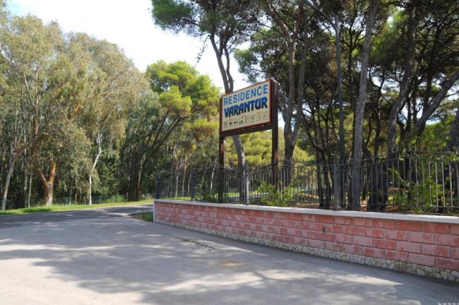 Ingresso del Residence Varantur con cartello, strada asfaltata e alberi intorno.