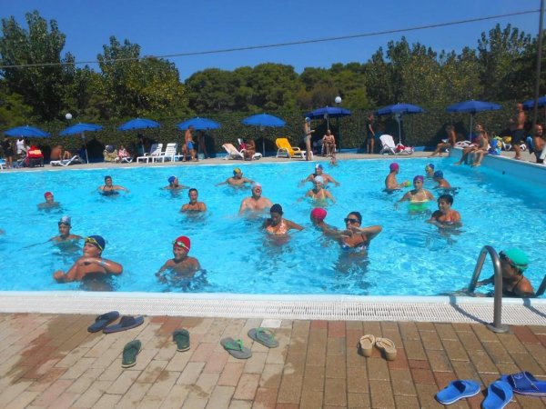 Persone nuotano in una piscina all’aperto, circondata da alberi e ombrelloni blu.