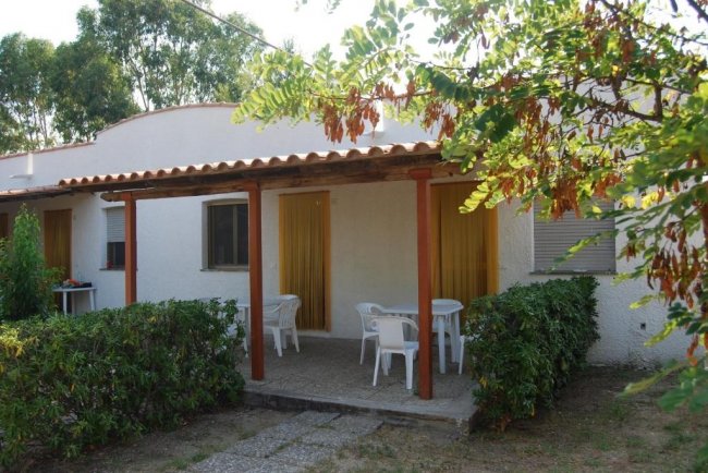 Una casetta bianca con veranda e tavolo, circondata da cespugli e alberi.