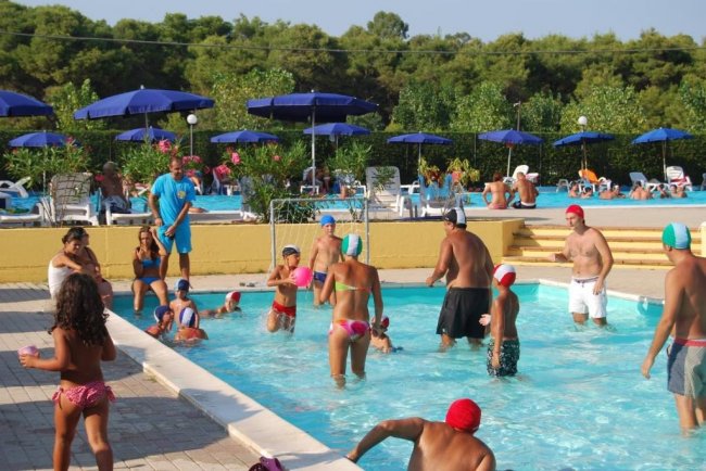 Gruppo di persone che giocano in piscina, con ombrelloni blu sullo sfondo.