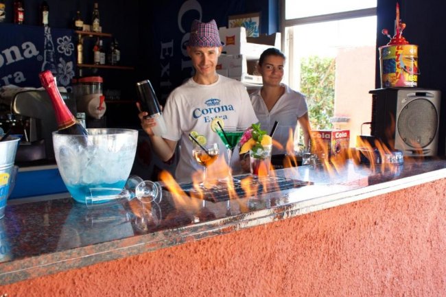Due persone dietro un bancone con fiamme sulla superficie e cocktail colorati in primo piano.