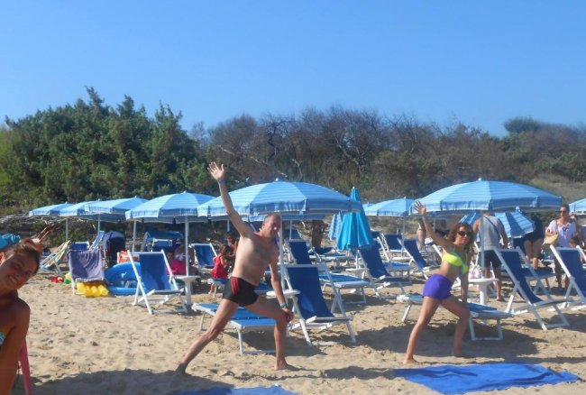 Persone che fanno esercizi di stretching su una spiaggia con sdraio blu e ombrelloni.