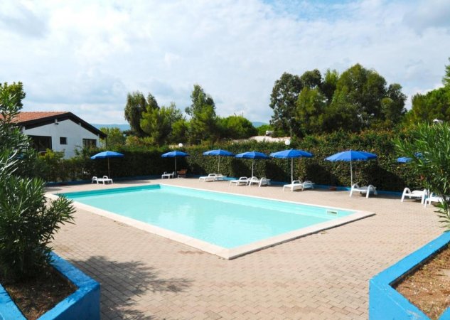 Una piscina rettangolare con sdraio e ombrelloni blu in un'area pavimentata circondata da alberi.
