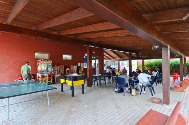 Un'area coperta con tavolini, sedie, calcio balilla e ping-pong, persone sedute e giochi.