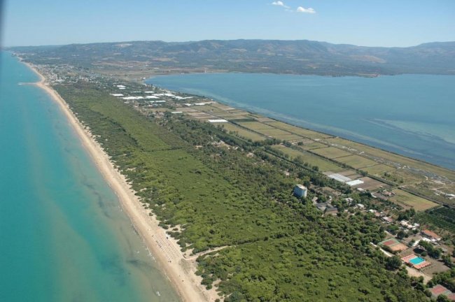 Vista aerea di una lunga spiaggia con acqua turchese, vegetazione e un lago.
